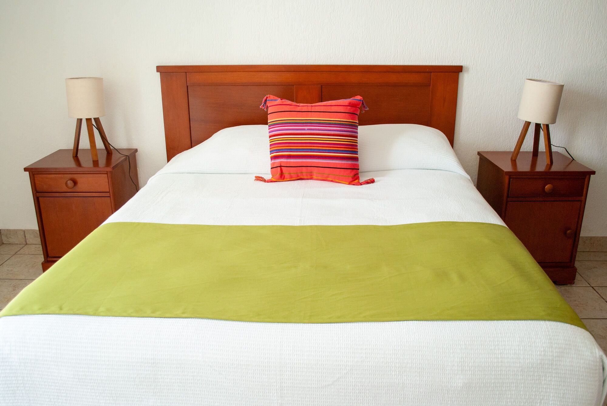 Een netjes opgemaakt bed met een groene deken, een gestreept kussen en twee nachtkastjes met lampen.