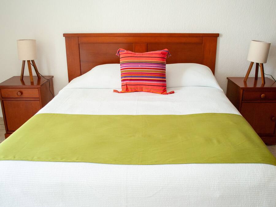 Een netjes opgemaakt bed met een groene deken, een gestreept kussen en twee nachtkastjes met lampen.