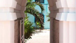Un pasillo estrecho con columnas blancas que conduce a un edificio azul con una ventana y palmeras.