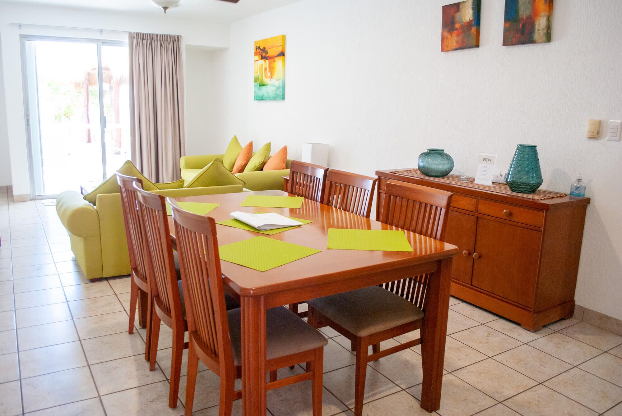 Un coin repas avec une table en bois, des sets de table verts et des chaises en bois. On aperçoit, à l’arrière-plan, un canapé jaune avec des coussins orange. Un buffet en bois, orné de vases décoratifs, est adossé au mur.