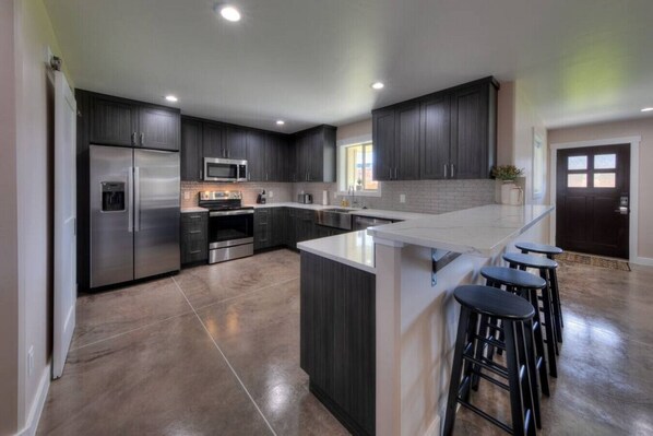A modern kitchen with dark cabinetry, stainless steel appliances, a large island with bar stools, and a tiled backsplash.