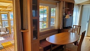 Un cuarto acogedor con un banco de madera, una mesa redonda y sillas. Hay una ventana con cortinas, un televisor y una repisa con objetos.