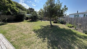 Un patio trasero con terraza de madera, un árbol y una casa de fachada blanca y contraventanas verdes.