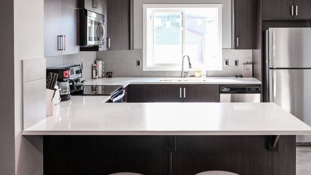 A modern kitchen with dark cabinetry, a white countertop, a stainless steel refrigerator, and a window providing natural light.
