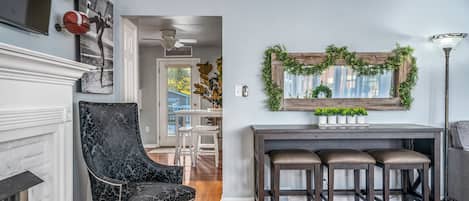 Una sala de estar con chimenea, un sillón negro, una mesa de centro de madera y tres taburetes, y un espejo con plantas.