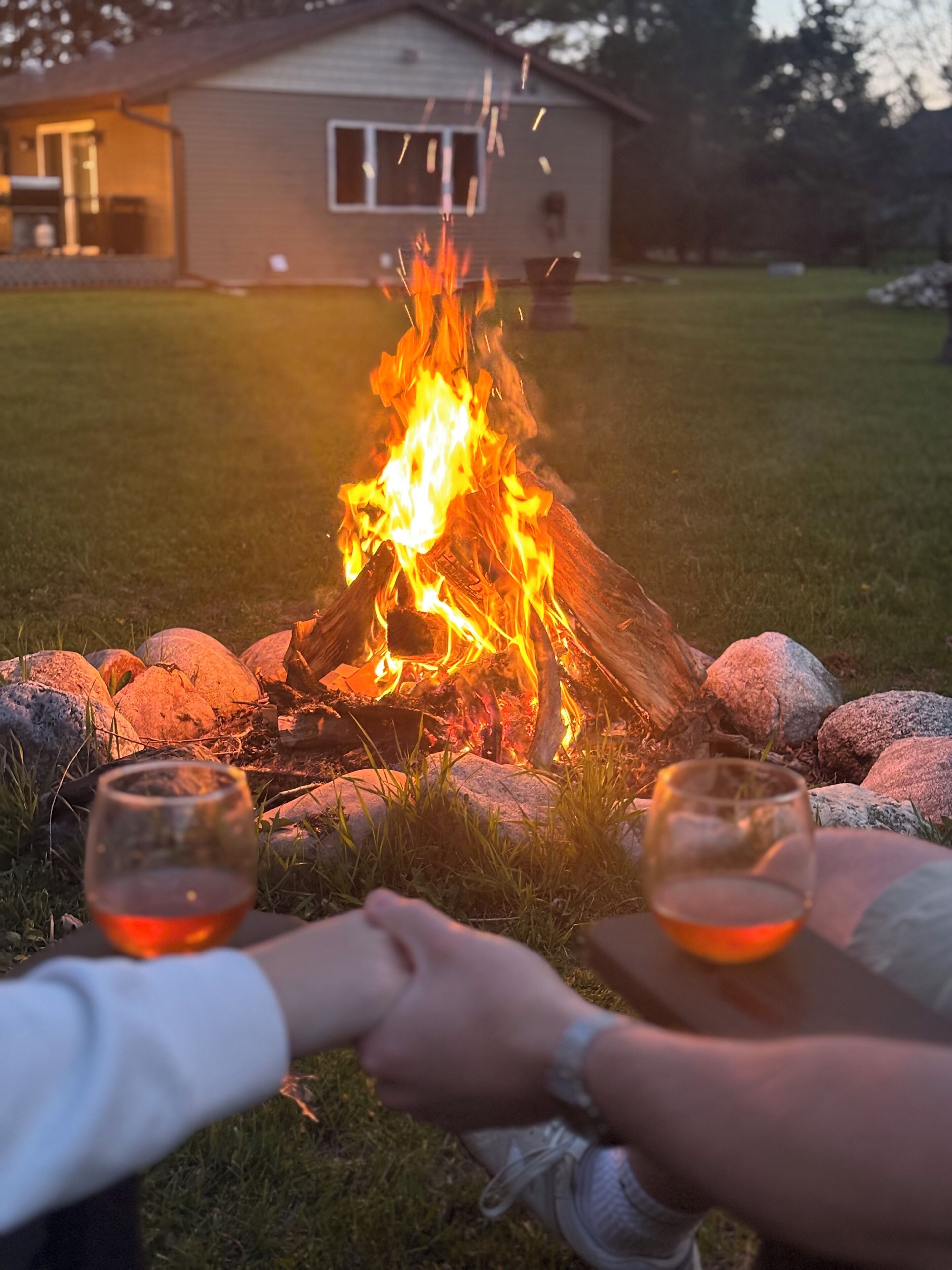 A campfire with two glasses of red beverage, surrounded by rocks, in a grassy area with a house in the background.