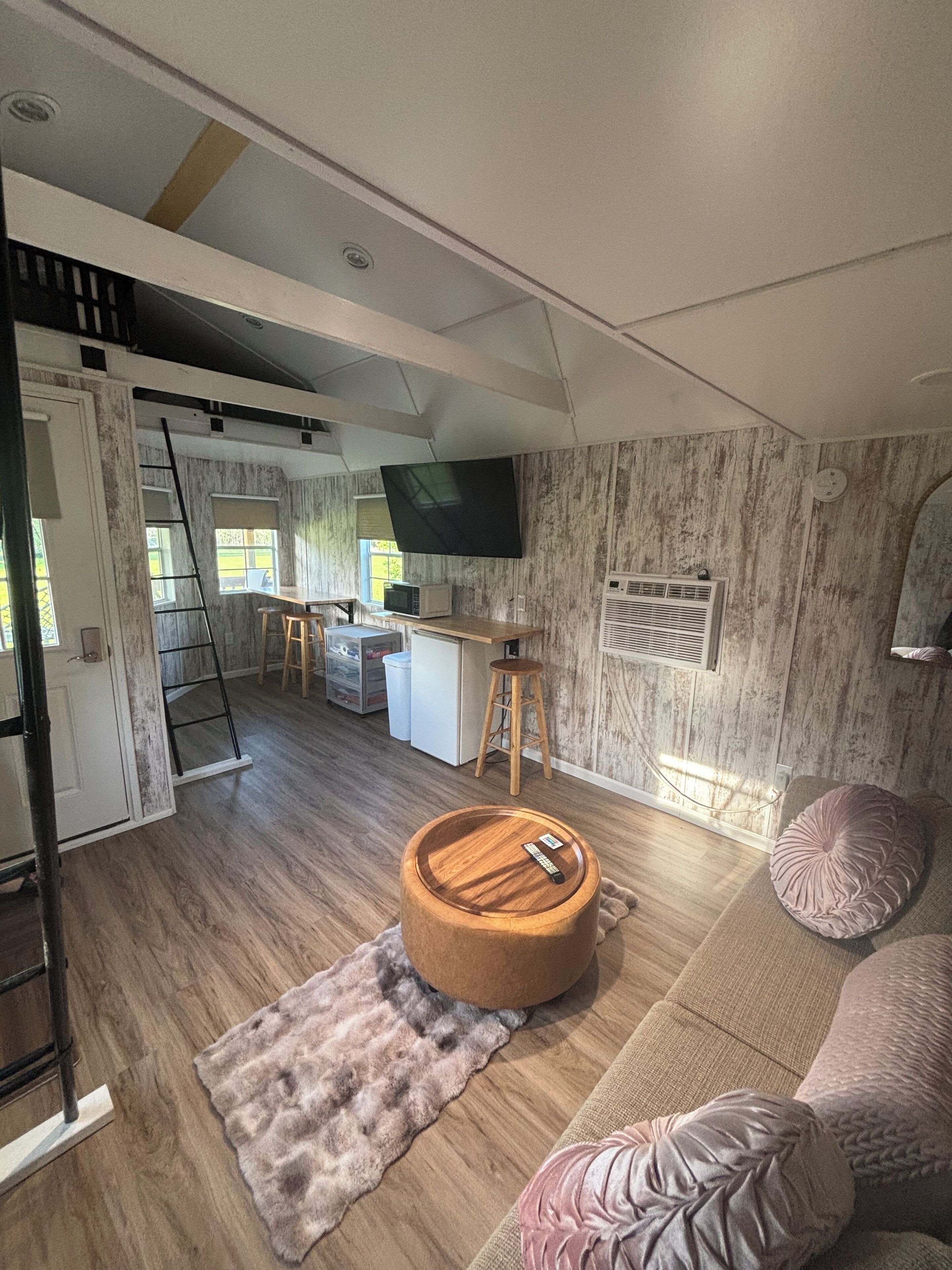 Una sala moderna con piso de madera, un otoman redonda de madera, un sofá color beige y un cojín rosa. Se observa una zona de cocina con un microondas y un taburete.