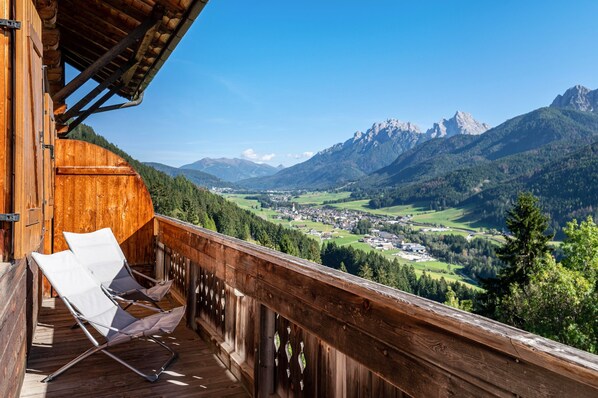 Un balcon en bois avec deux chaises blanches, donnant sur un paysage montagneux avec un village en contrebas.