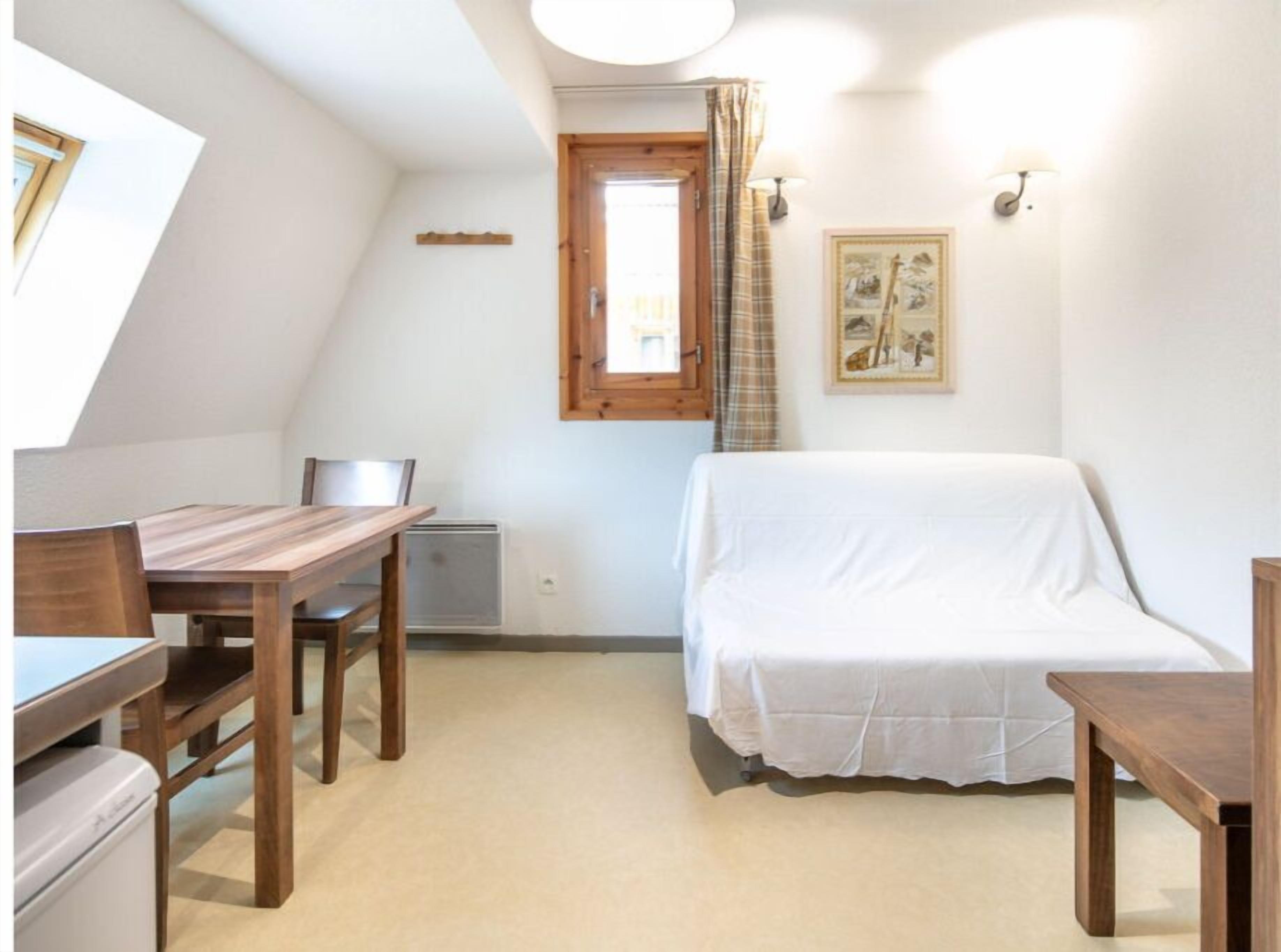 Une petite pièce agréable et chaleureuse, avec une table et des chaises en bois, un lit recouvert d’une courtepointe blanche, une fenêtre avec des rideaux et une lampe fixée au mur.