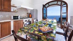 A dining area with a table set for a meal, kitchen appliances, and a view of the sea. - Casa Sofia 310 steps (Positano)
