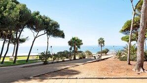 Een geplaveid pad, omzoomd door bomen en banken, dat naar een strand leidt met de zee op de achtergrond.