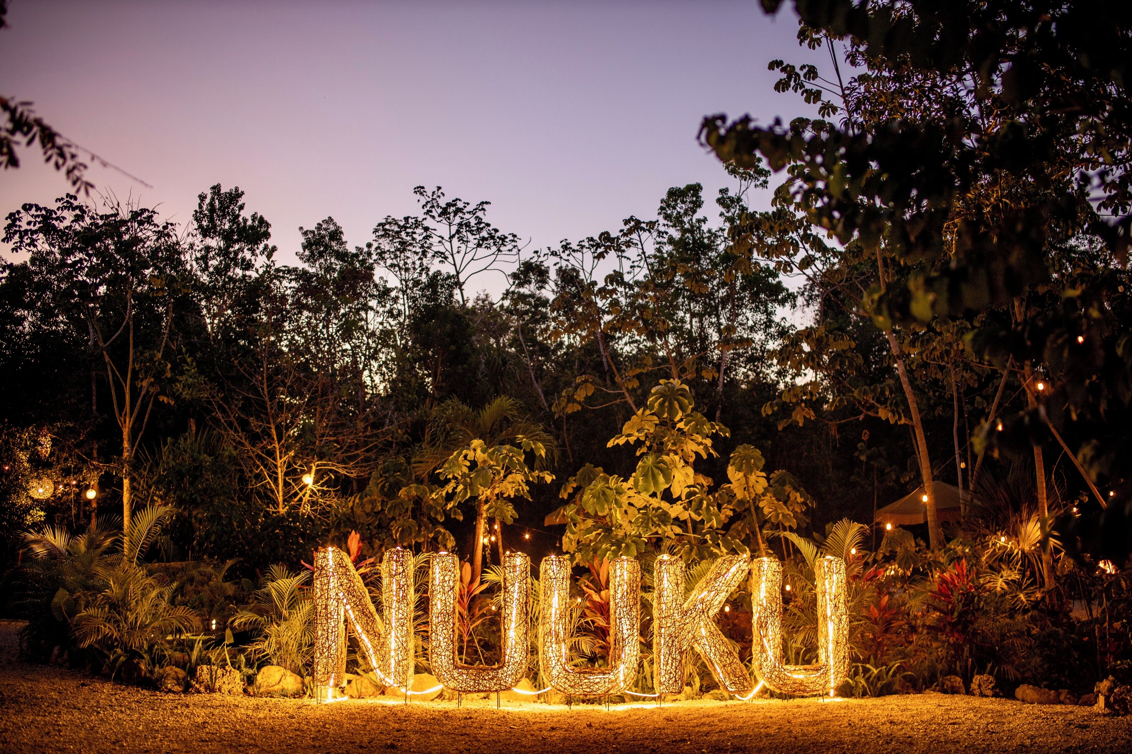 Illuminated letters spelling 'NILUKU' in a garden at dusk.