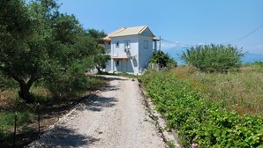 Un bâtiment blanc avec un toit recouvert de tuiles, un chemin de gravier menant à l’entrée et une végétation luxuriante de part et d’autre.
