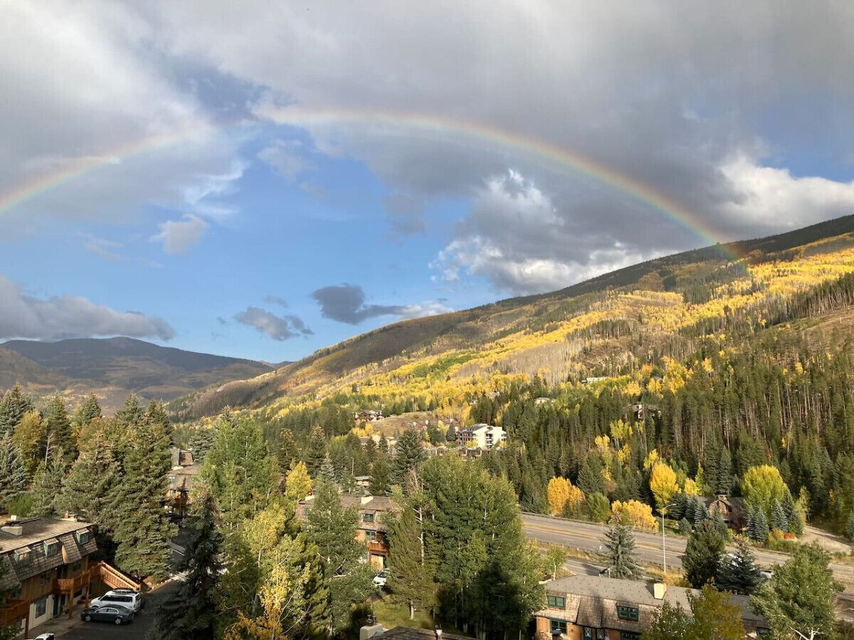 Eine Berglandschaft mit Herbstlaub, ein Regenbogen und eine Straße, die zu einer Häufung von Häusern führt.