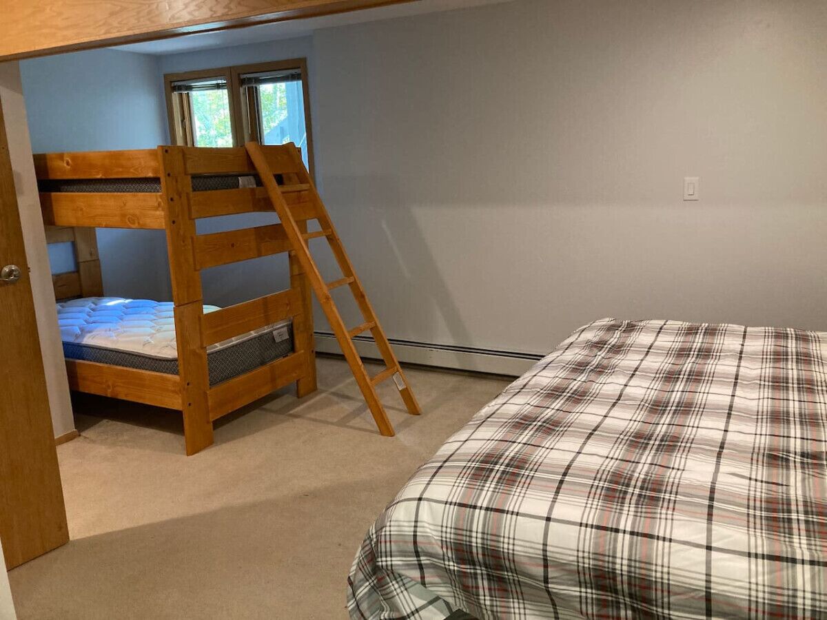 A bedroom with a bunk bed, a ladder, a plaid bedspread, and a window with a view.