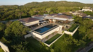 Una casa moderna de varios pisos con una gran piscina, rodeada de exuberante vegetación y colinas al fondo.