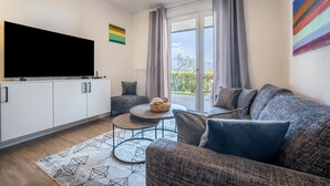 Ein modernes Wohnzimmer mit Flachbildfernseher, einer Couch, einem Couchtisch und Blick ins Freie durch eine Glastür.