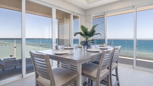 Een eetgedeelte met een tafel gedekt voor zes personen, grote ramen met uitzicht op het strand en de oceaan, en een potplant op de tafel.