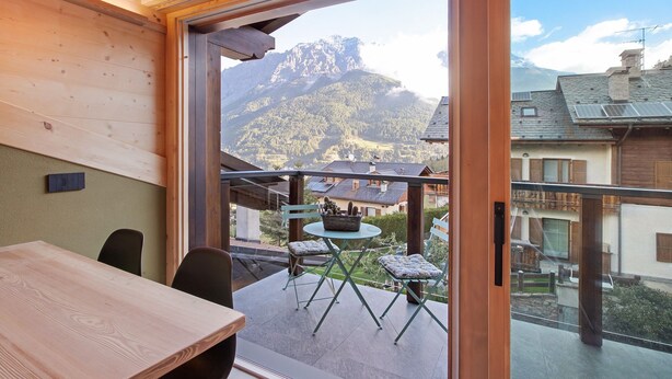 A room with a wooden ceiling, a balcony with a table and chairs, and a view of mountains.