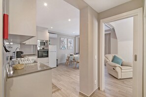 Een moderne keuken met een magnetron, oven en gootsteen, een eethoek met tafel en stoelen, en een woonkamer met een bank en een deur naar een andere kamer.