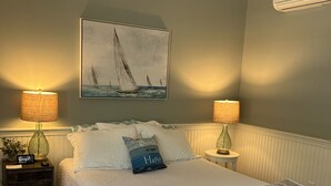 A bedroom with a bed, two bedside lamps, a ceiling fan, an air conditioning unit, and a painting of sailboats.