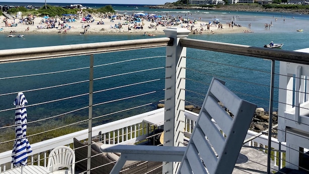 A balcony with white chairs and a striped umbrella, overlooking a crowded beach with people and boats.