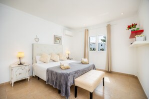 A bedroom with a bed, bedside tables, a bench, a window, and a shelf with flowers.