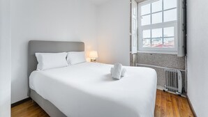 A neatly made bed with white linens and pillows, a bedside lamp, and a window with a view of buildings. - GuestReady - Modern Duplex, Neo-Classical Building (Porto)