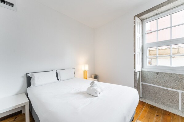 A minimalist hotel room with a large bed, a bedside table, a lamp, and a window with a view. - GuestReady - Modern Duplex, Neo-Classical Building (Porto)