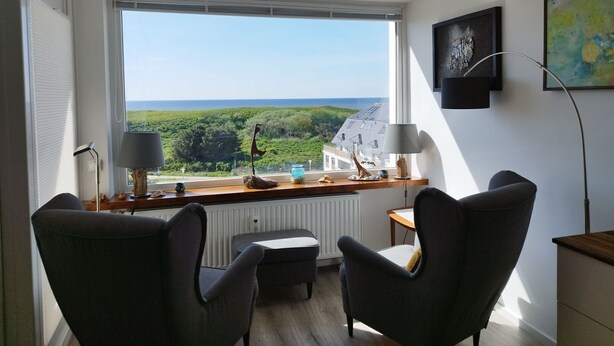 A modern room with a large window offering a view of greenery and a building. There are two armchairs, a small table, and a floor lamp. - House Brandenburg App. 43 with lake view - 2 room apartment with breathtaking views (Sylt)