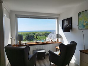 A modern room with a large window offering a view of greenery and a building. There are two armchairs, a small table, and a floor lamp.