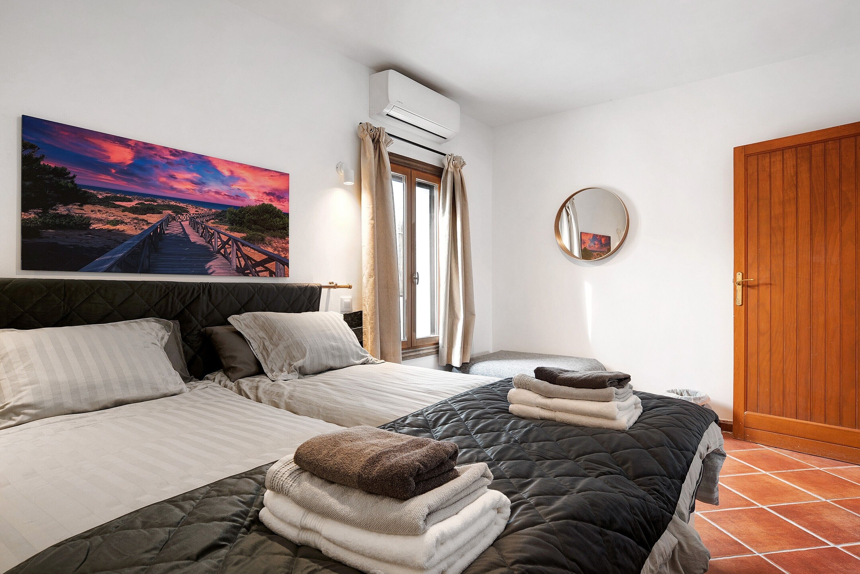A bedroom with two beds, a large painting, an air conditioning unit, a mirror, and a wooden door.