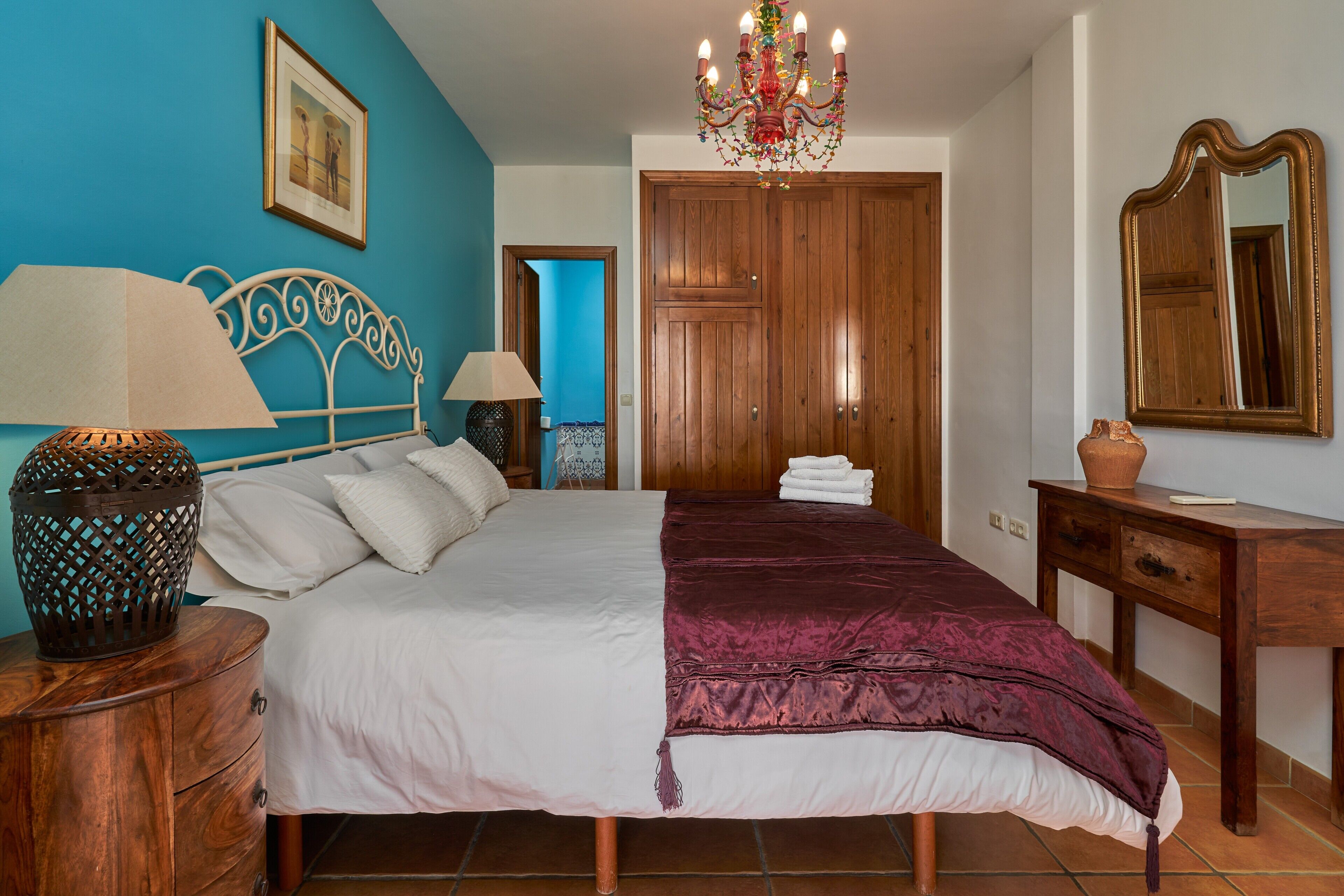 A bedroom with a bed, two bedside tables, a wooden wardrobe, a mirror, and a chandelier.