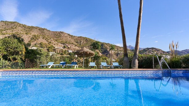 A swimming pool with a patterned edge, lounge chairs, palm trees, and a mountainous backdrop.