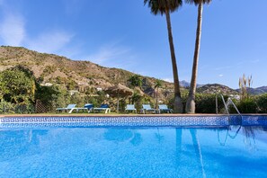A swimming pool with a patterned edge, lounge chairs, palm trees, and a mountainous backdrop.