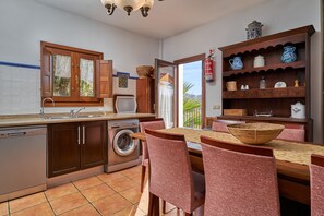 A kitchen with a washing machine, a dining table with chairs, and a shelf with kitchenware.
