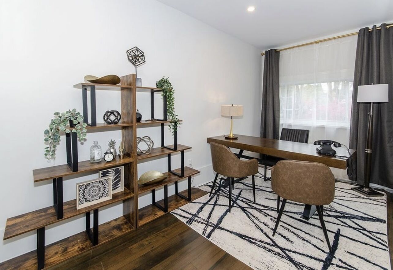 Een moderne woonkamer met een houten boekenkast, een eettafel met stoelen, een lamp en een groot raam met gordijnen.