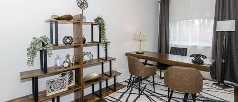 Een moderne woonkamer met een houten boekenkast, een eettafel met stoelen, een lamp en een groot raam met gordijnen.