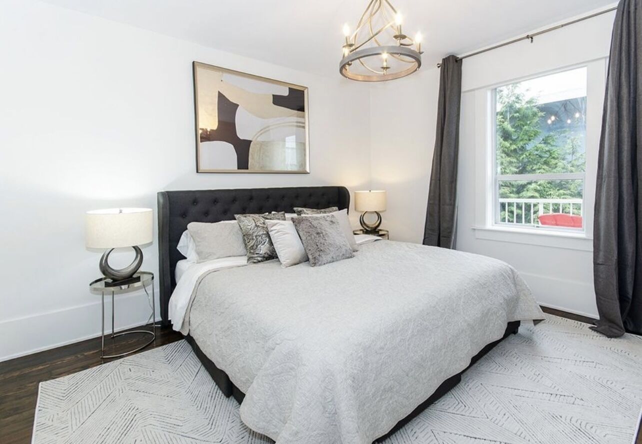 Quarto moderno com uma cama grande, duas luminárias de cabeceira, um lustre, uma obra de arte abstrata emoldurada e uma janela com cortinas.
