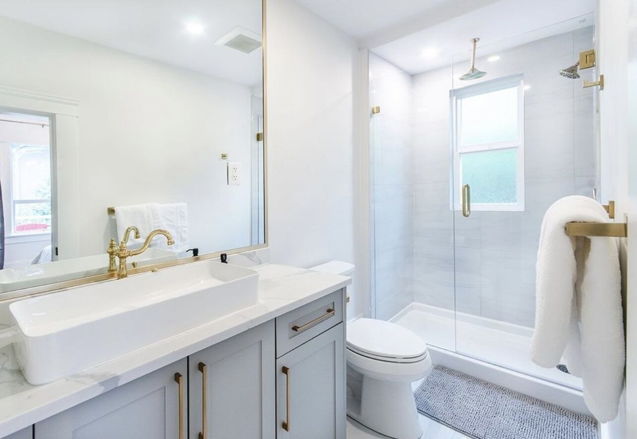 Een moderne badkamer met een groot wit wastafelmeubel, gouden kranen, een toilet en een douche met glazen wand.