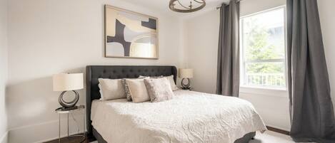 Un dormitorio con una cama grande, dos lámparas de noche, un candelabro, una ventana con cortinas y un cuadro abstracto colgado en la pared.