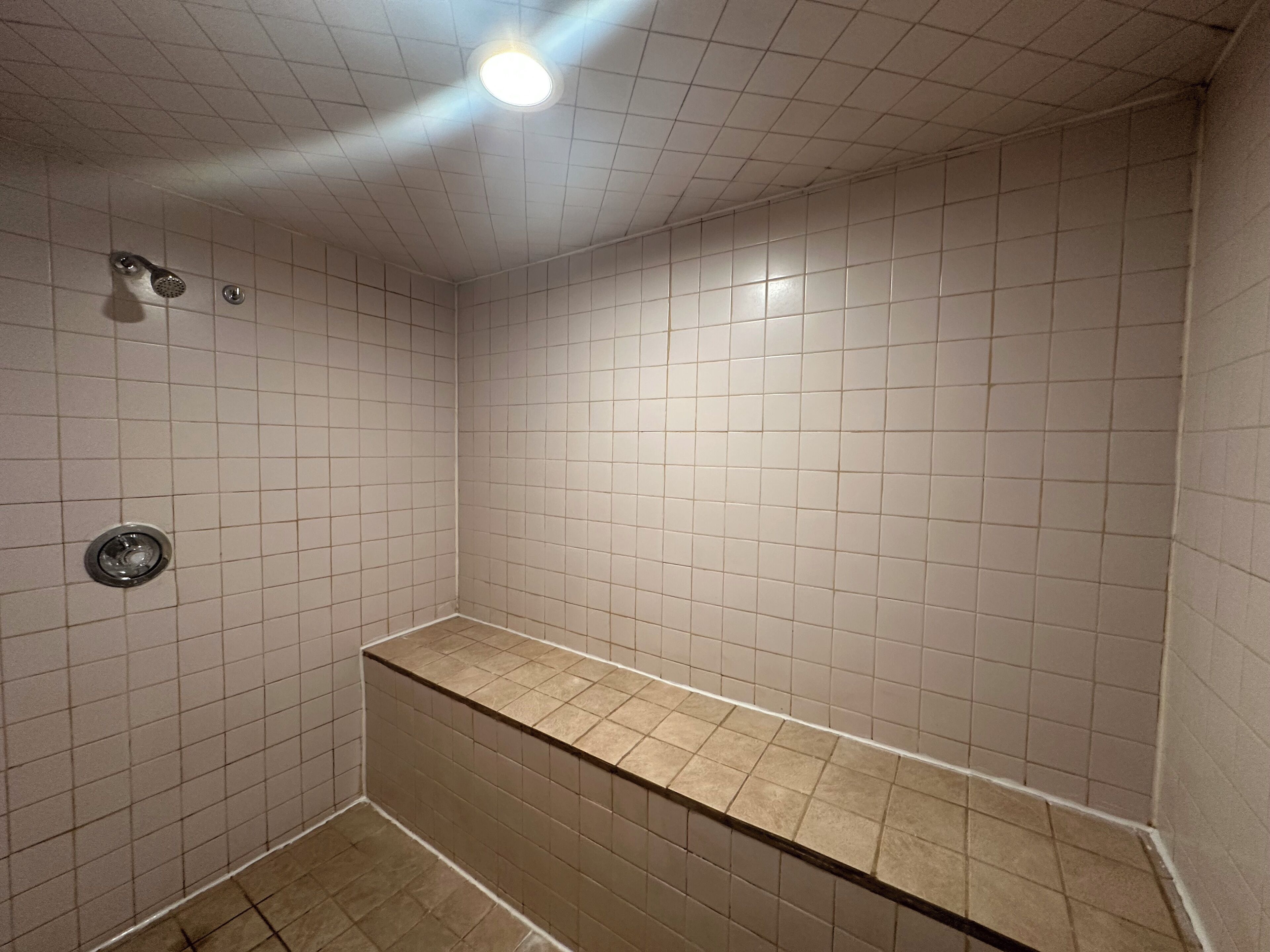 Une petite douche carrelée, avec un banc, un luminaire au plafond et un drain.