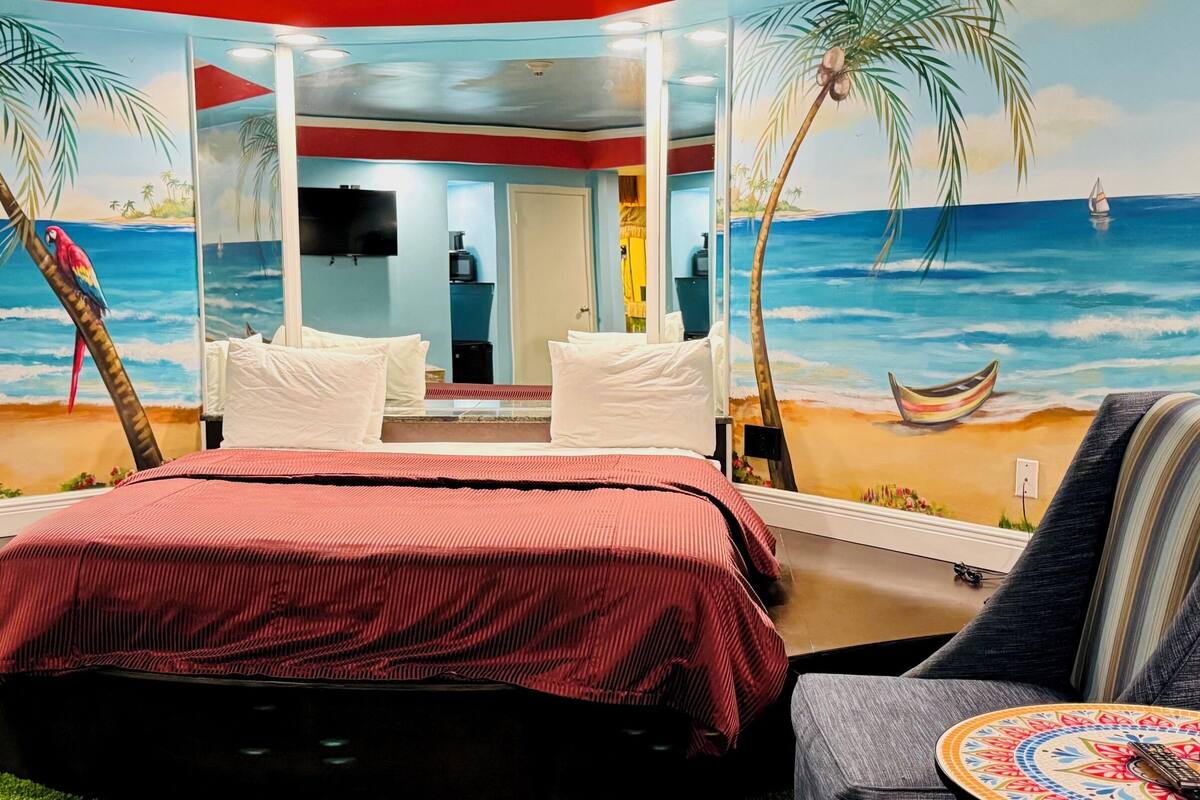 Une chambre avec une fresque murale représentant une plage, un lit recouvert d’une couverture rouge, une chaise et une petite table sur laquelle est posée une assiette décorative.