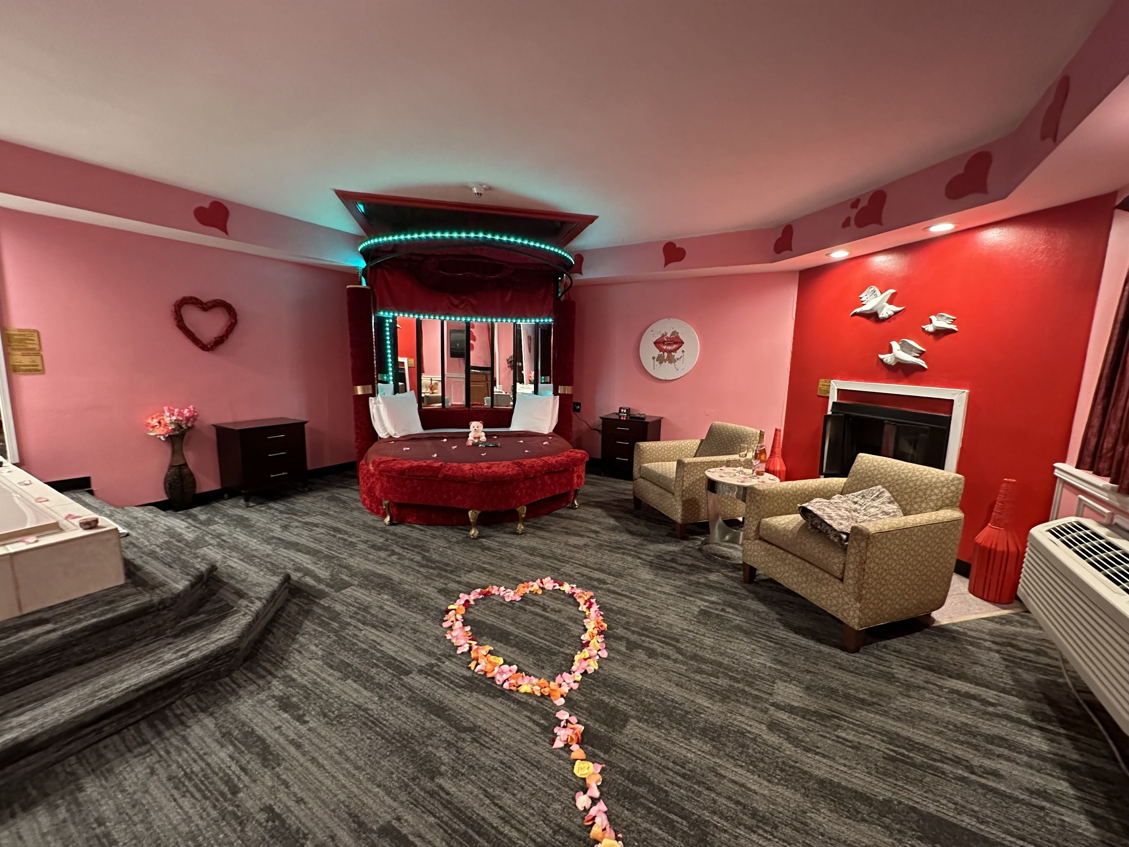 Una habitación con alfombra roja, un arreglo floral con forma de corazón, una cama tapizada en terciopelo rojo, chimenea y un sofá.