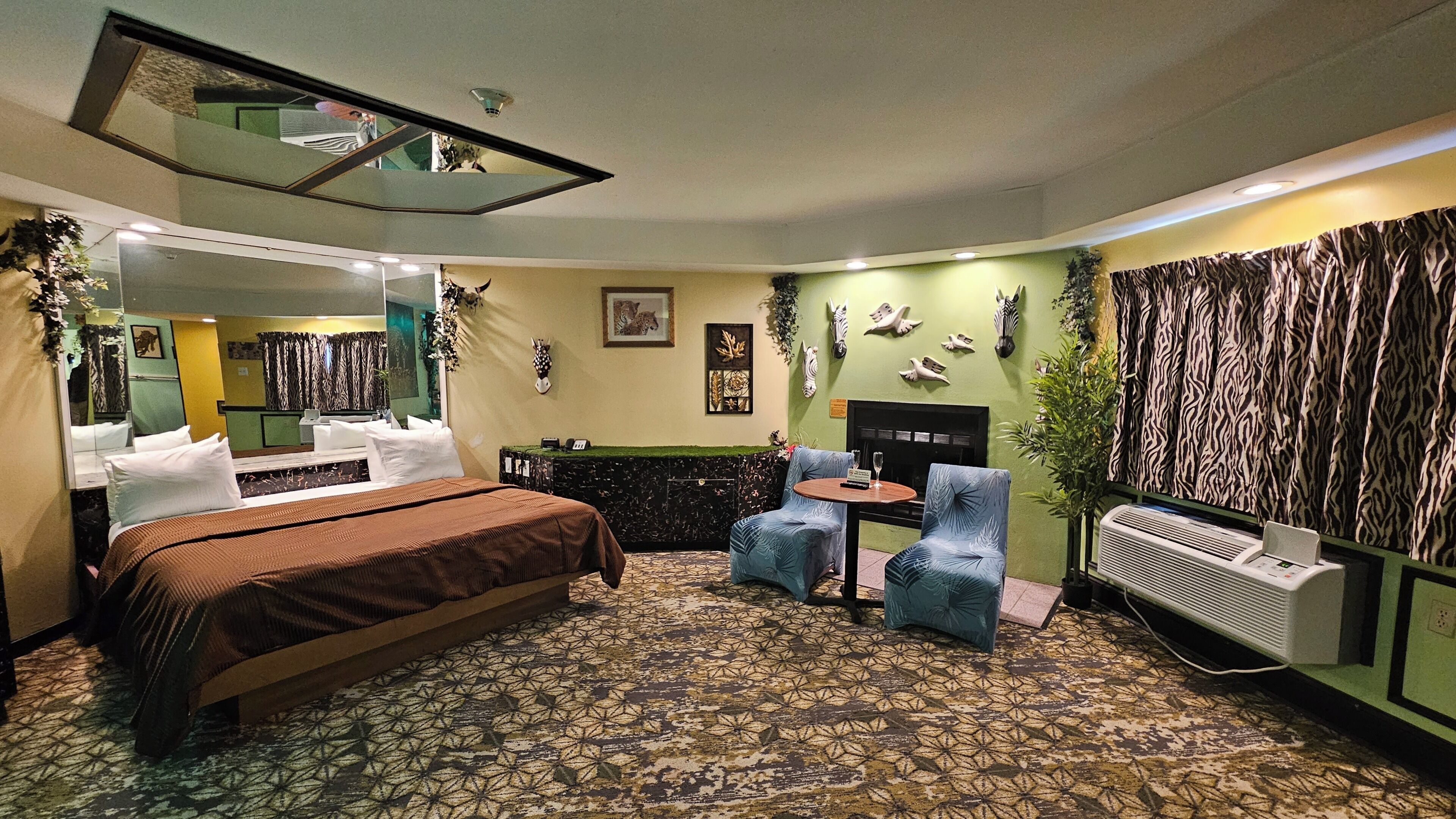 A hotel room with a bed, two chairs, a small table, a mirror, a fireplace, and a wall-mounted air conditioner.