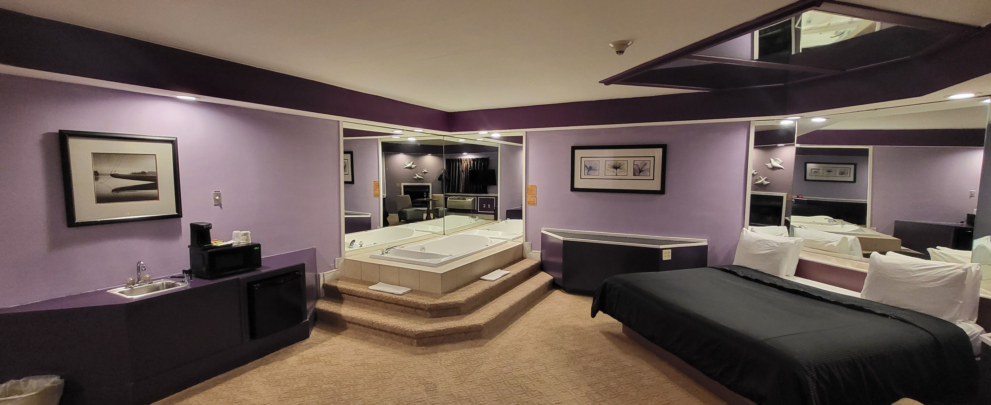 A room with a bed, a small desk, a vanity with a mirror, a bathtub, and a large mirror reflecting the room.
