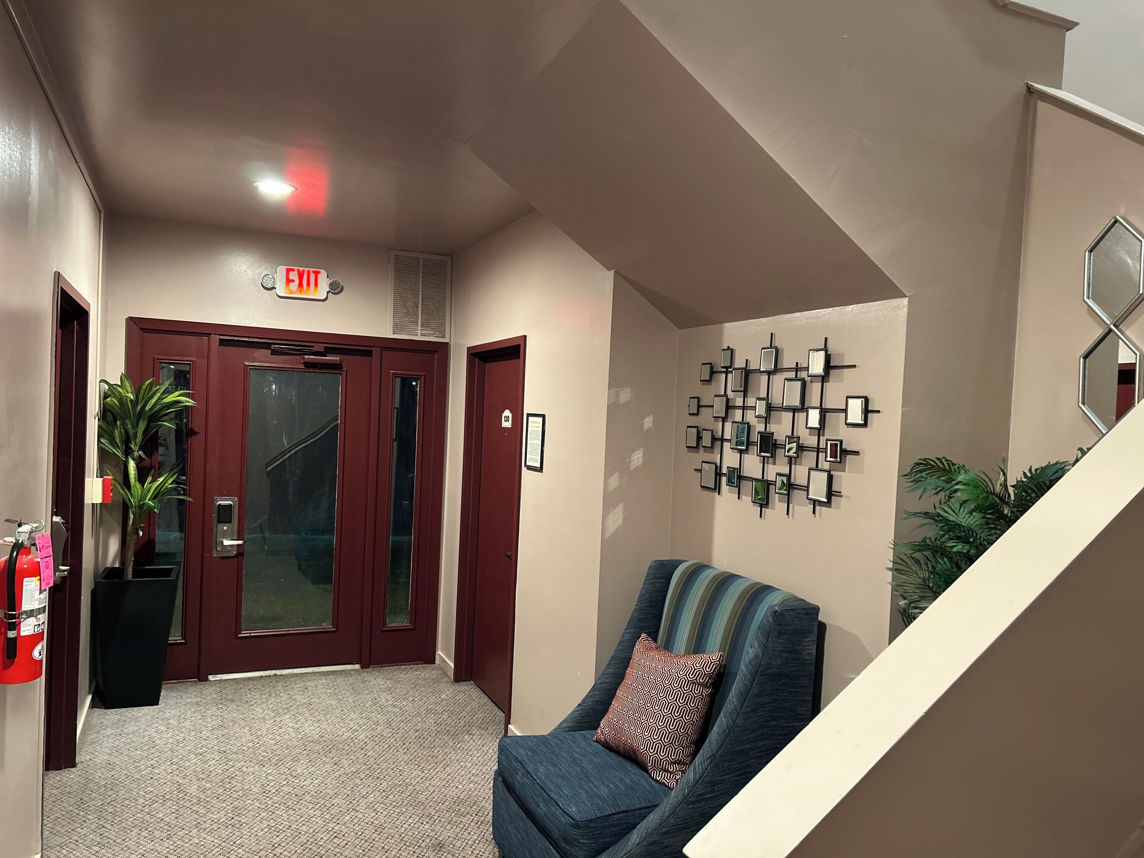A hotel lobby with a red door marked "EXIT," a blue chair with a patterned pillow, a wall-mounted mirror, and a staircase.
