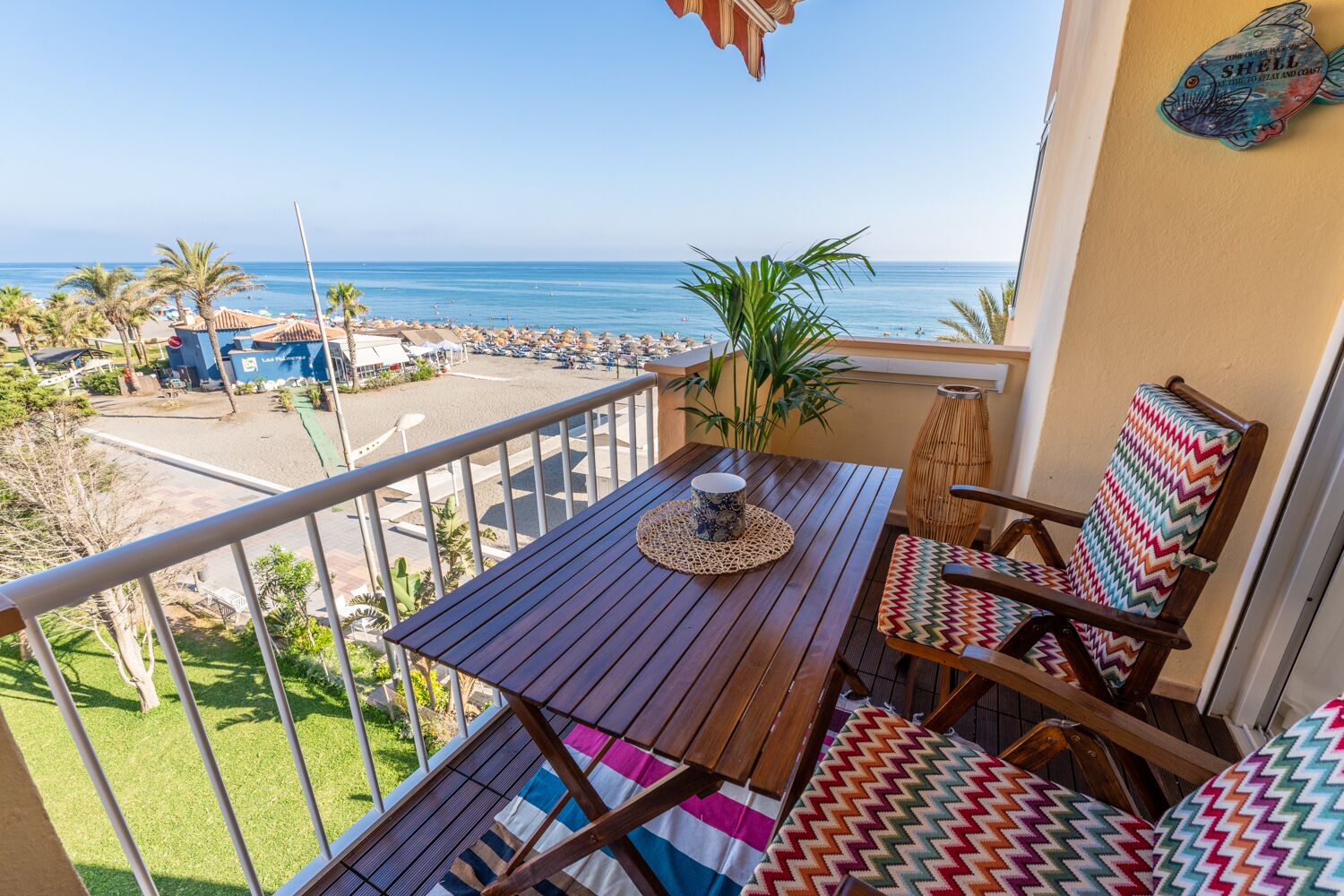 A balcony with a wooden table, two colorful chairs, a potted plant, and a view of the beach and ocean.