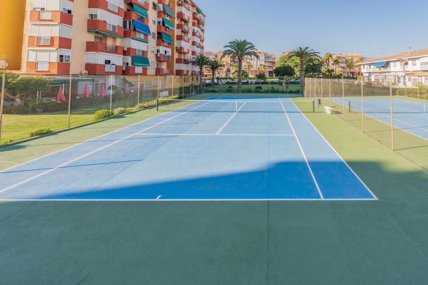 En udendørs tennisbane omgivet af et hegn, med en boligbygning i baggrunden.