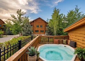 A wooden cabin with a hot tub and a fenced deck, surrounded by trees and a clear sky.